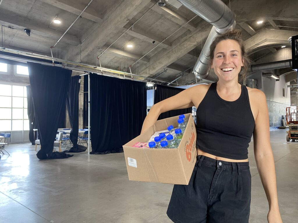 Une femme tient à la hanche une boite rempli de denrées diverse, dans un hangar aménagé en loge